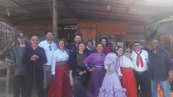   Integrantes do grupo de danças “Inclusão Sem Fronteiras” sorriem para a foto; eles trajam roupas do tradicionalismo gaúcho, na maioria, mulheres com longos vestidos de prenda. 